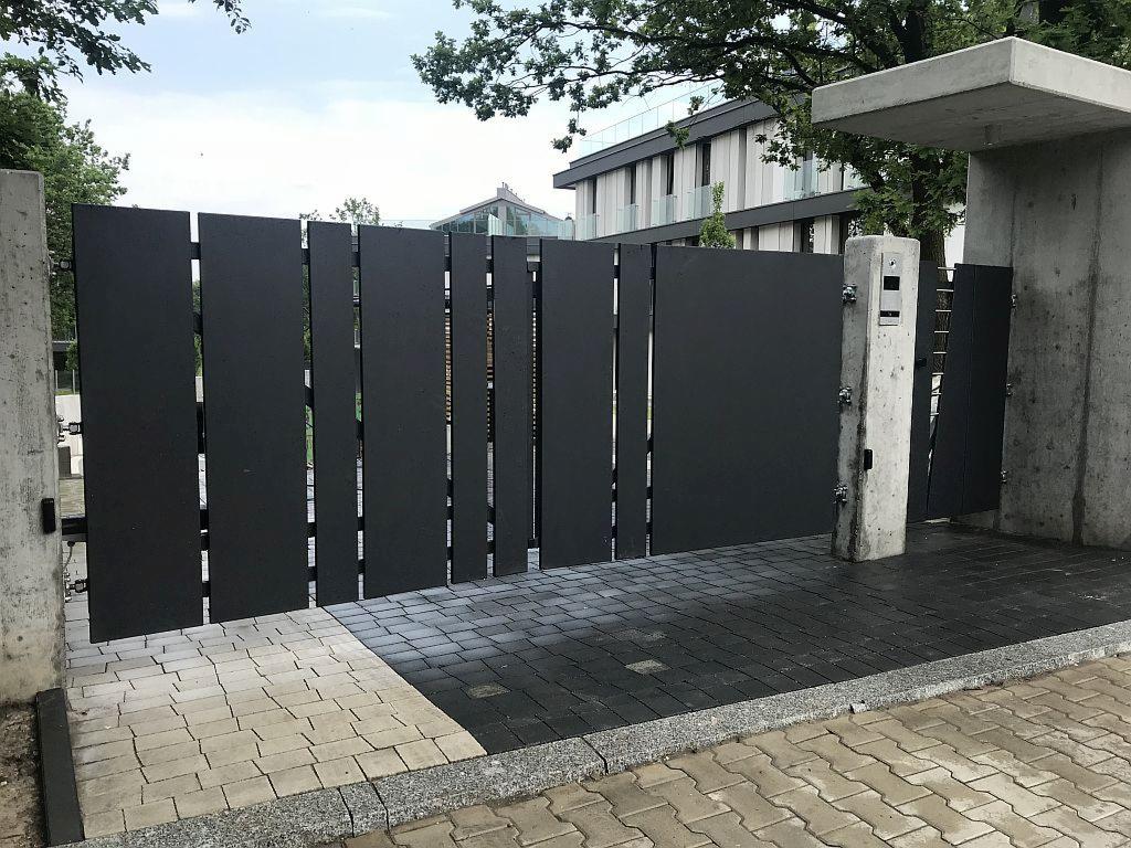 Nowoczesna, grafitowa brama wjazdowa z pionowych paneli, osadzona na betonowych słupach, z widocznym fragmentem kostki brukowej w dwóch odcieniach szarości i budynkiem w tle.