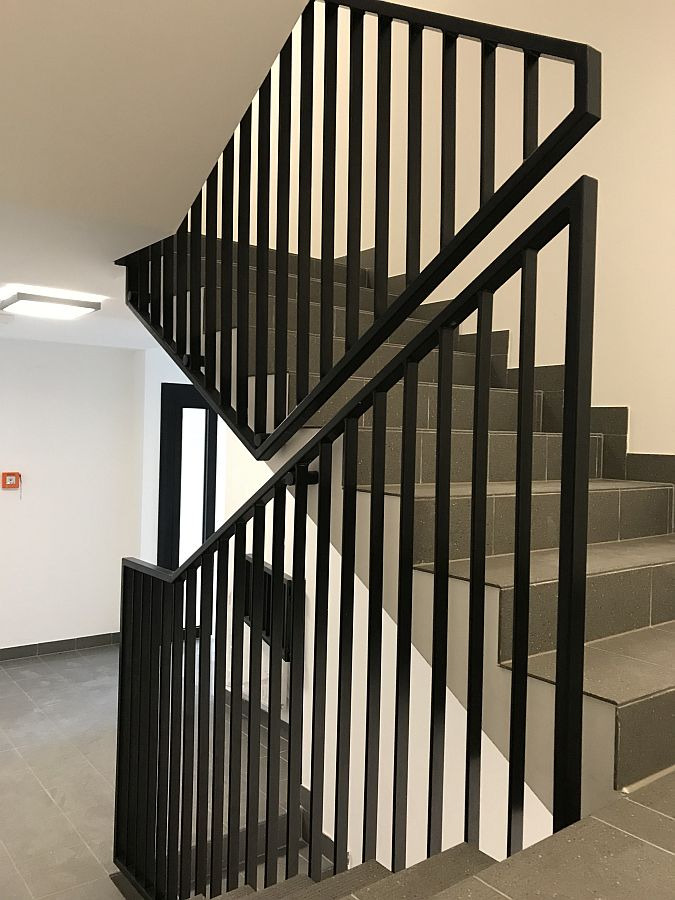 Wnętrze klatki schodowej z nowoczesną, czarną balustradą ze stali i szarymi, kwadratowymi płytkami na schodach i podłodze.