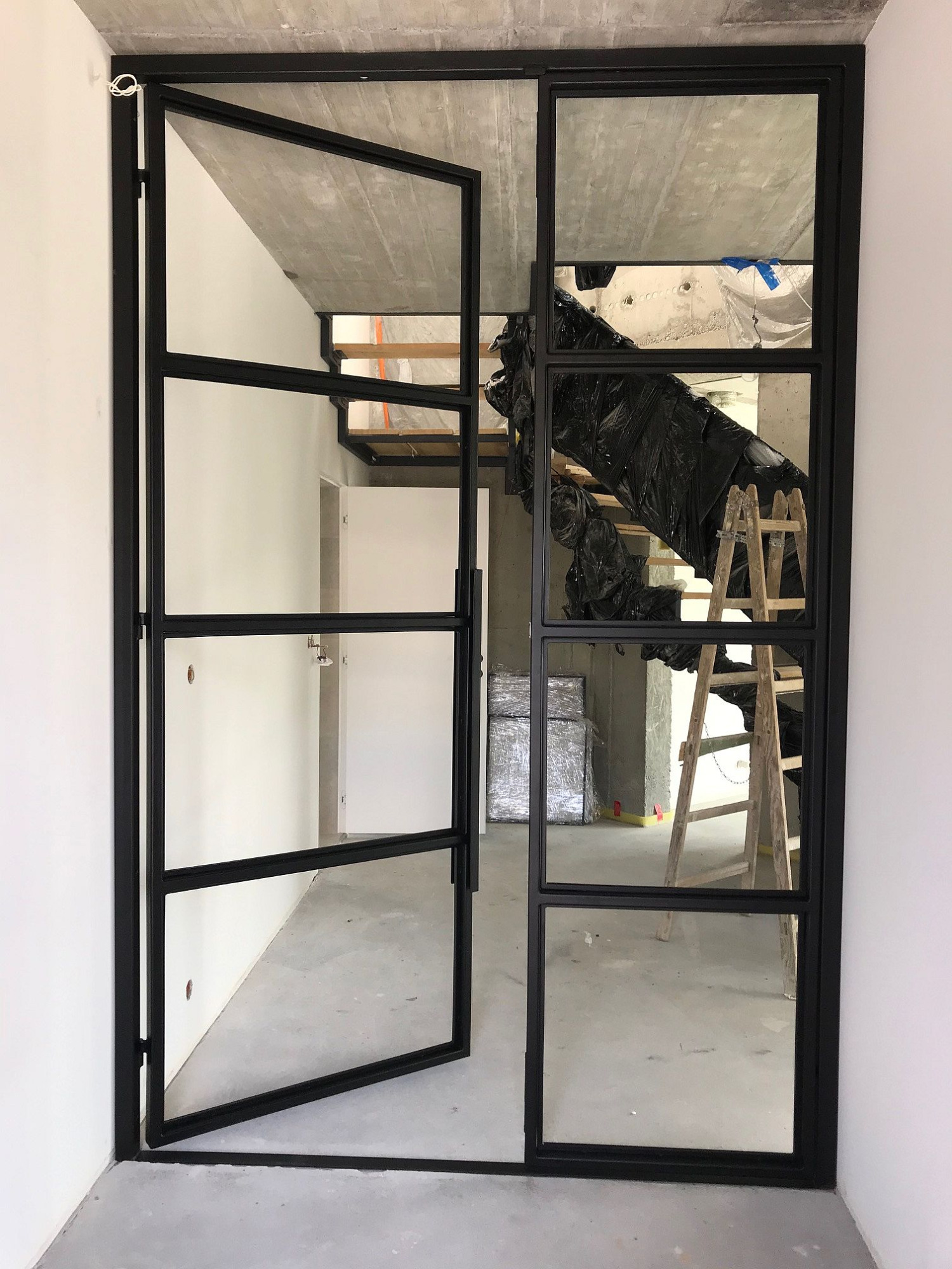 Nowoczesne, przeszklone drzwi stalowe z czarnymi szprosami, częściowo otwarte, widok na surowe wnętrze w trakcie remontu: betonowy strop, zabezpieczone schody i drabina.