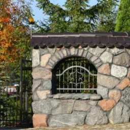 Kamienny mur z ozdobną kratą i daszkiem z dachówki bitumicznej, obok metalowa furtka i drzewa w tle. Solidne ogrodzenie posesji.