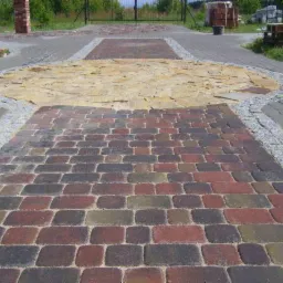 Wzorzysty podjazd z kostki brukowej i kamienia, z elementami dekoracyjnymi z drobnego granitu. Urozmaicona kolorystyka kostki, okrągła wstawka z jasnego kamienia.