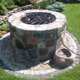 Kamienna studnia ogrodowa z dekoracyjnym metalowym elementem w środku, otoczona brukowanym kręgiem, na tle zielonego trawnika i roślinności.