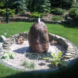 Ogród z fontanną z naturalnego kamienia otoczoną kamieniami ozdobnymi i roślinami, na tle zadbanego trawnika i dekoracyjnych nasadzeń.