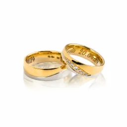 Dwa złoto-srebrne obrączki ślubne, jedna z rzędem brylantów, obie z wygrawerowanymi datami i imionami na wewnętrznej stronie, leżące na białym tle z odbiciem.