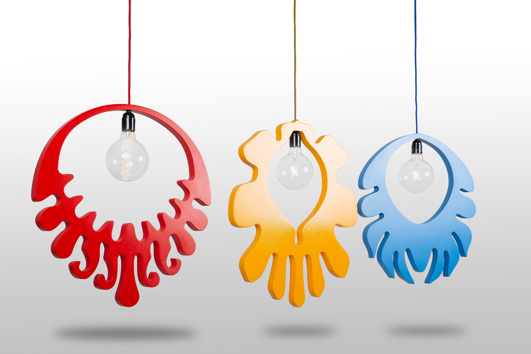 Trzy kolorowe, nowoczesne lampy wiszące z żarówkami Edison w geometrycznych, ażurowych obudowach: czerwona w kształcie rozgwiazdy, żółto-pomarańczowa z motywem liści i błękitna z falistymi elementami.