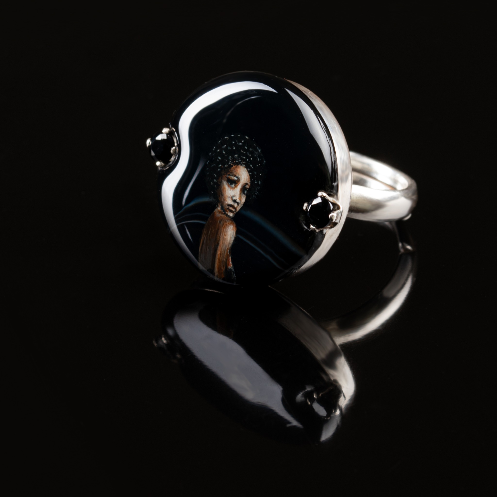 Pierścionek z dużym, okrągłym, czarnym oczkiem, na którym namalowany jest portret kobiety, oprawiony w srebro i ozdobiony małymi, czarnymi kamieniami, leżący na czarnym, błyszczącym tle.