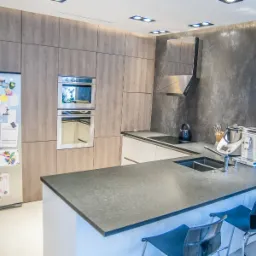 Nowoczesna kuchnia z drewnianymi frontami szafek, szarą ścianą, wyspą z ciemnym blatem i transparentnymi krzesłami barowymi, lodówka z magnesami.