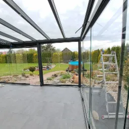 Szklana zabudowa tarasu z widokiem na ogród z basenem i placem zabaw, drabina oparta o szybę.