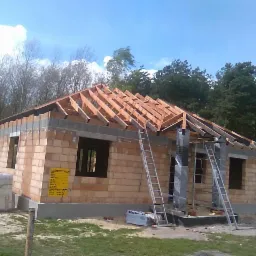 Surowy budynek jednorodzinny w trakcie budowy, z widoczną konstrukcją drewnianą dachu bez pokrycia, dwie drabiny opierają się o ścianę, a na ścianie wisi żółta tablica informacyjna.