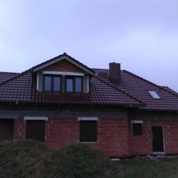 Ciesielstwo-Dekarstwo - Dom w trakcie budowy z nowym, bordowym dachem z dachówki ceramicznej, widoczne okno dachowe i komin, ściany z czerwonej cegły, teren wokół domu częściowo zagospodarowany.