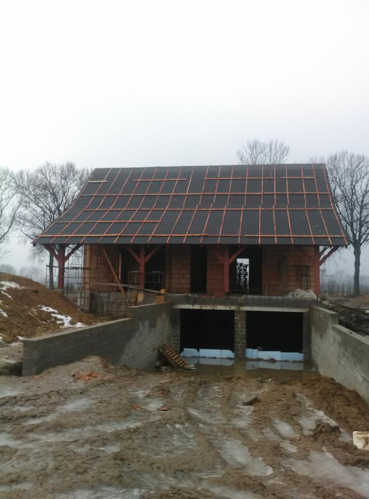 Budowa dachu na budynku gospodarczym w trakcie realizacji: widoczne deskowanie, izolacja i więźba dachowa, na tle pochmurnego nieba i błotnistego terenu.