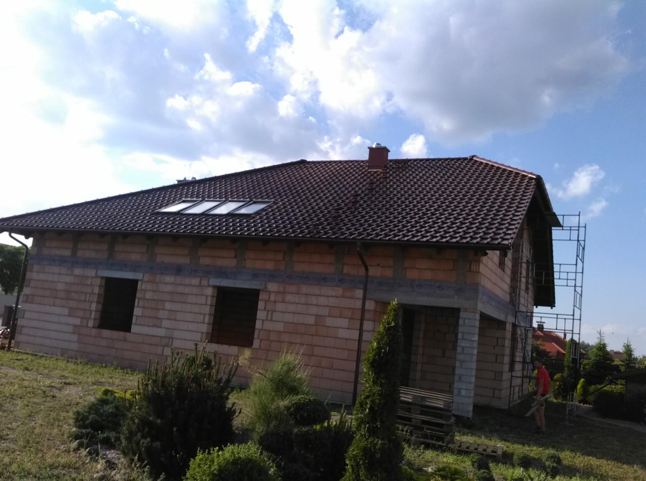 Dom w trakcie budowy z nowym dachem z brązowej dachówki ceramicznej i oknami dachowymi, widoczne rusztowanie i elementy wykończeniowe.