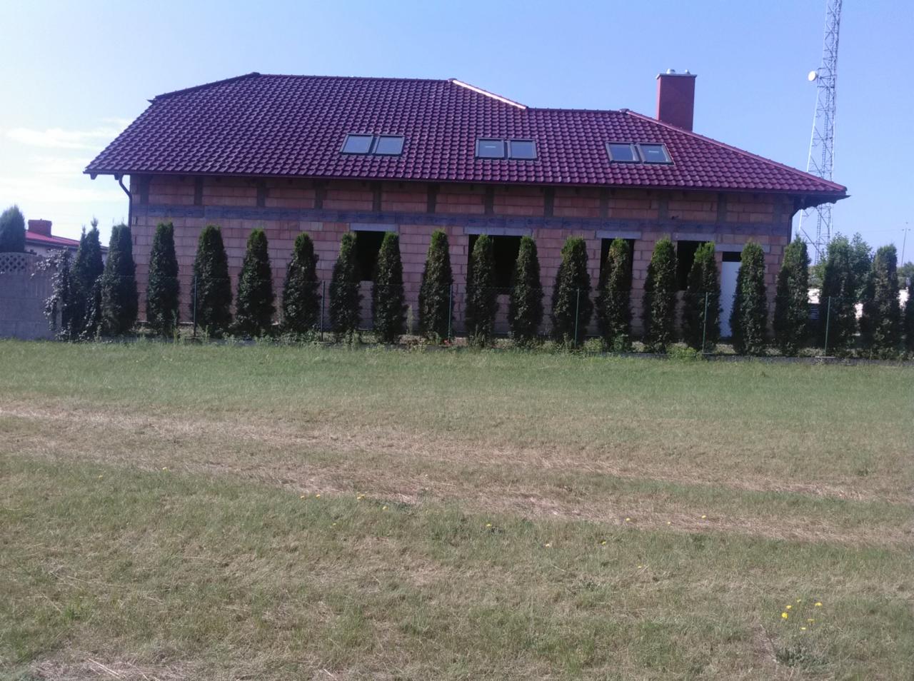 Niedokończony budynek z czerwoną dachówką i oknami dachowymi, widok z zewnątrz na duży trawnik i rząd drzew iglastych.