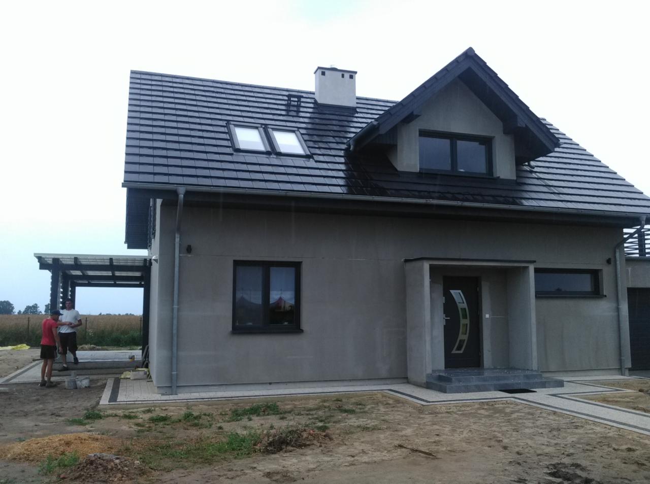 Nowoczesny dom jednorodzinny z ciemną dachówką karpiówką i oknami dachowymi, widoczne rynny i obróbki blacharskie, fragment pergoli z boku domu, dwaj mężczyźni w tle.