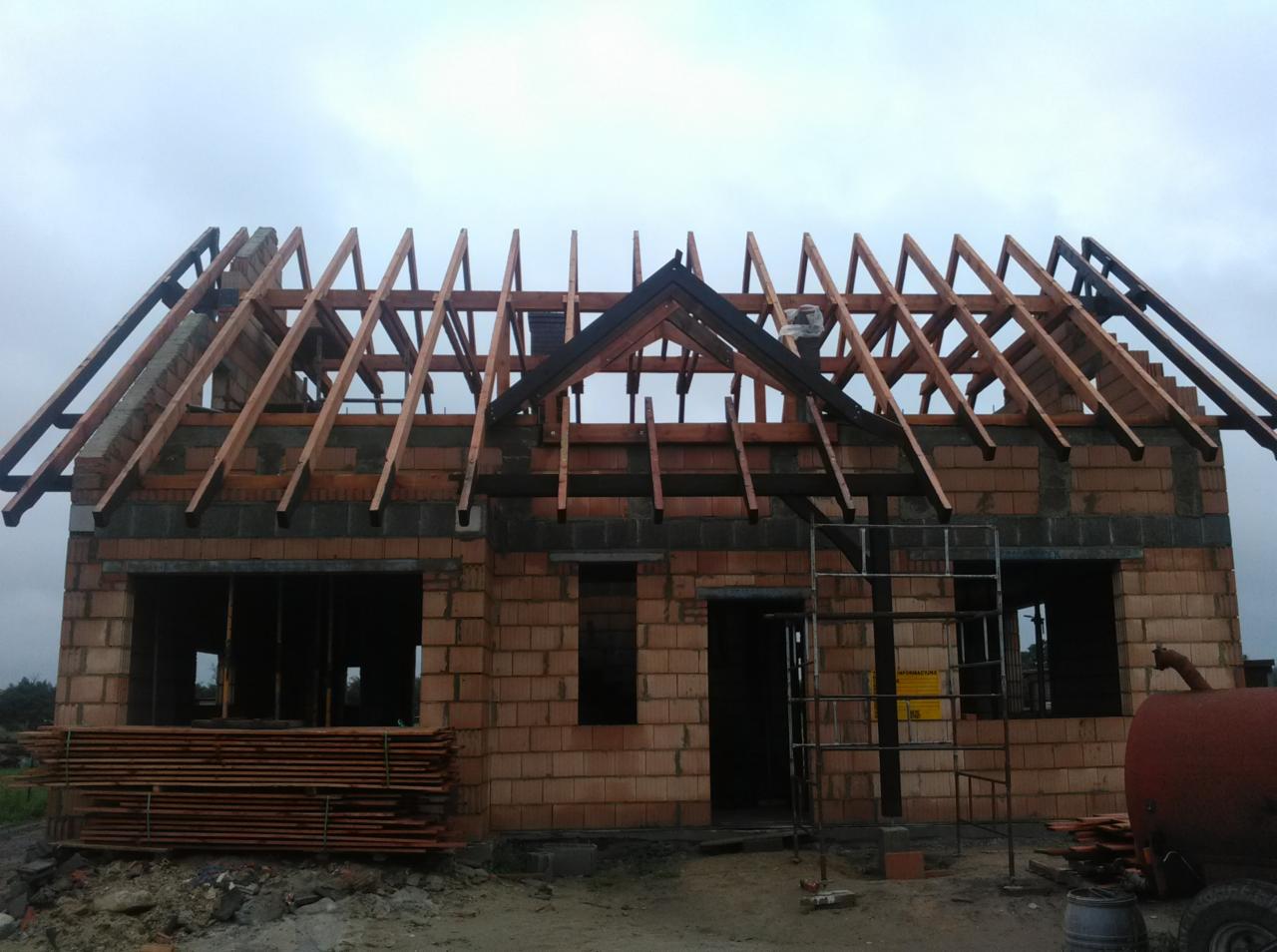 Budowa domu jednorodzinnego w trakcie montażu drewnianej konstrukcji dachu, widoczne elementy więźby dachowej i murłat, w tle pochmurne niebo.