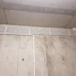 Równoległe białe rury w korytarzu, przymocowane do surowej betonowej ściany, z widoczną puszką instalacyjną w rogu.