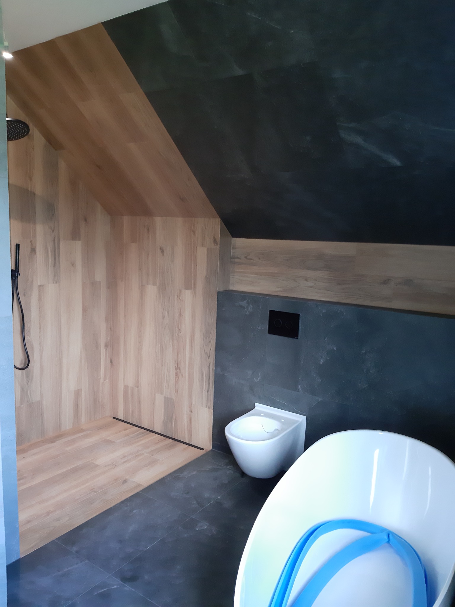 Nowoczesna łazienka w skosie z drewnopodobną okładziną prysznica, grafitowymi ścianami i podłogą oraz białą armaturą.