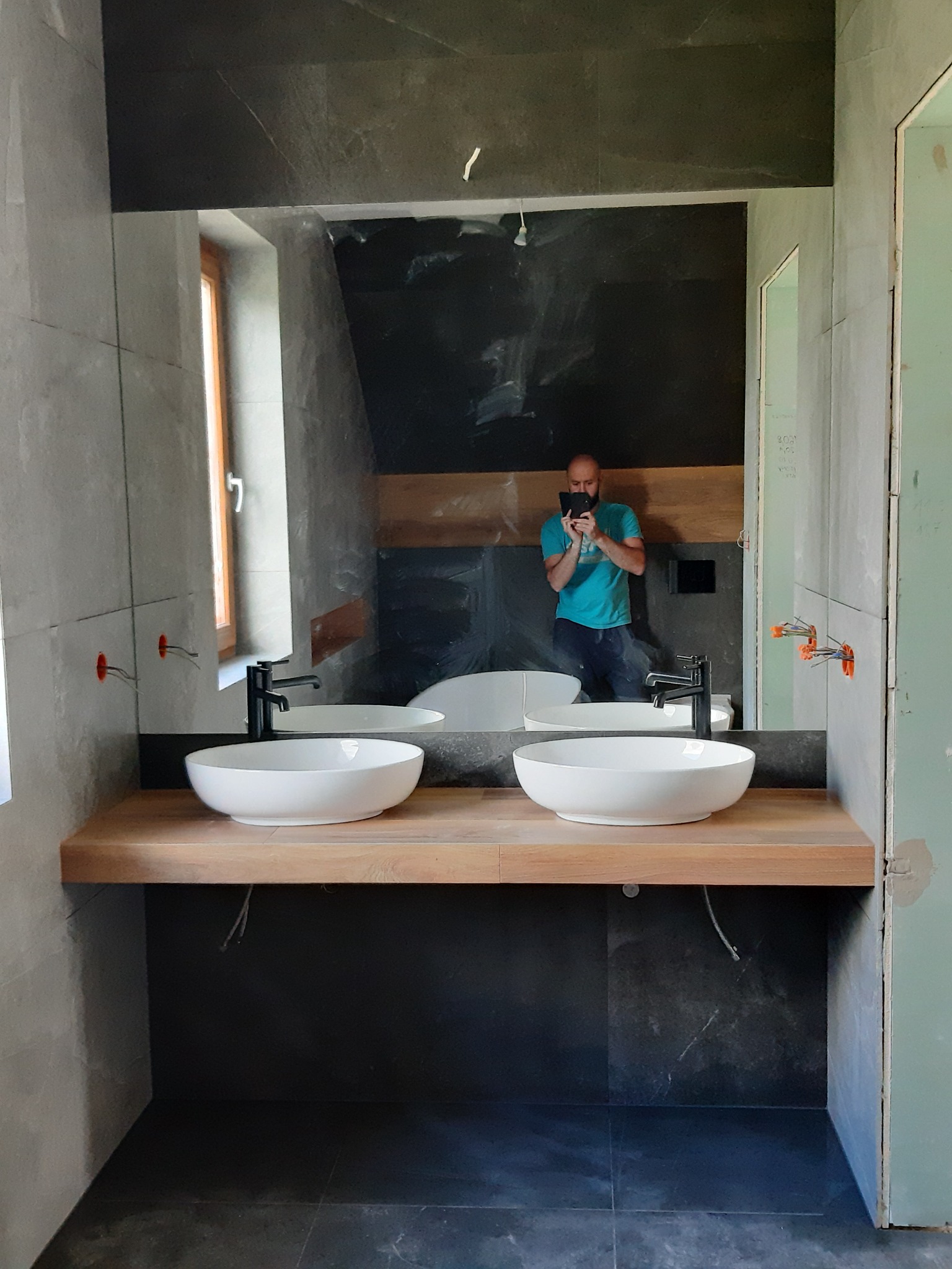 Nowoczesna łazienka w trakcie wykańczania, z dwiema białymi umywalkami na drewnianym blacie, duże lustro odbijające osobę robiącą zdjęcie, widoczne szare płytki na ścianach i podłodze...