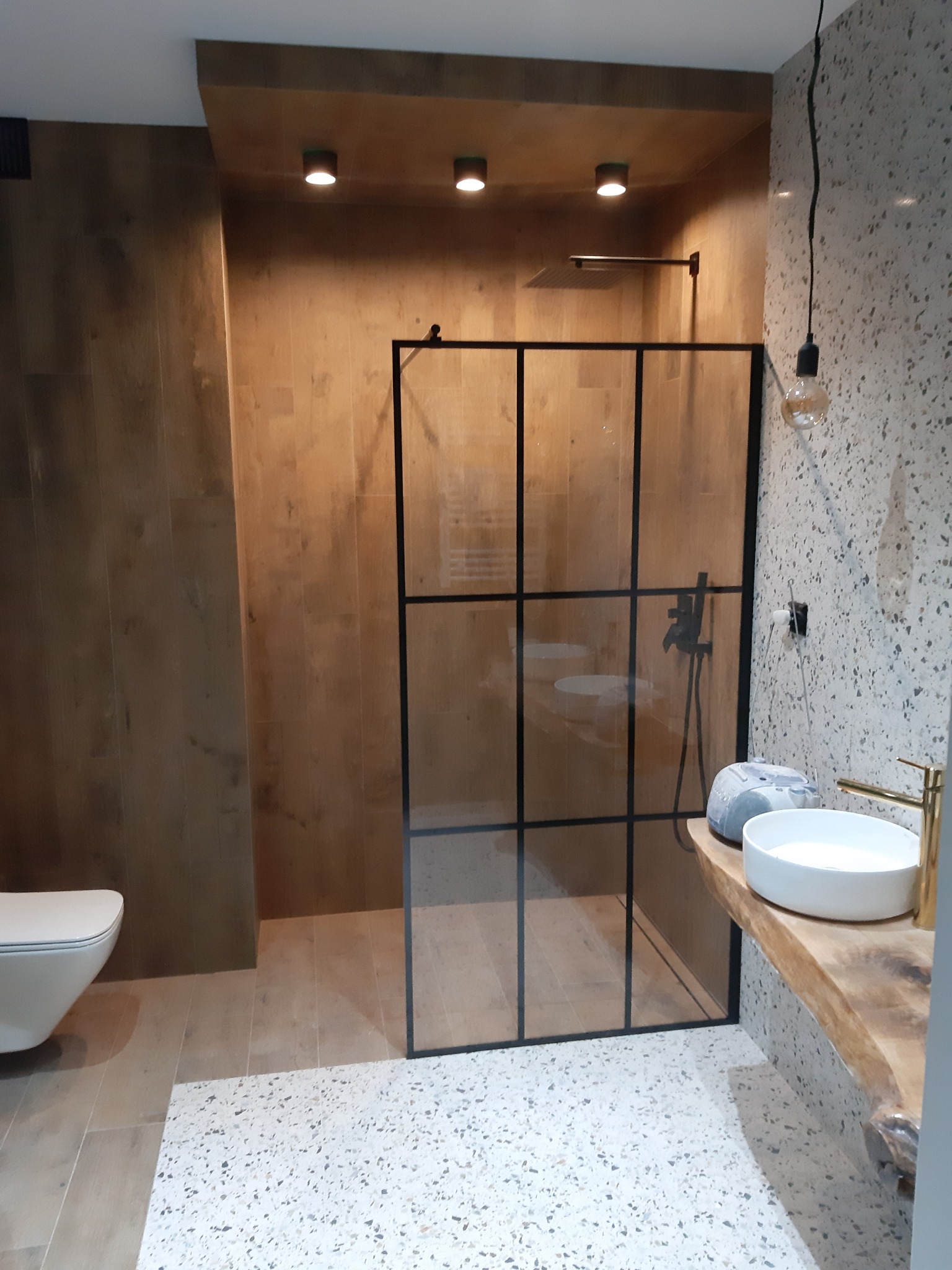 Nowoczesna łazienka z kabiną prysznicową z czarnymi ramami, umywalką nablatową na drewnianym blacie i podłogą z lastryko.