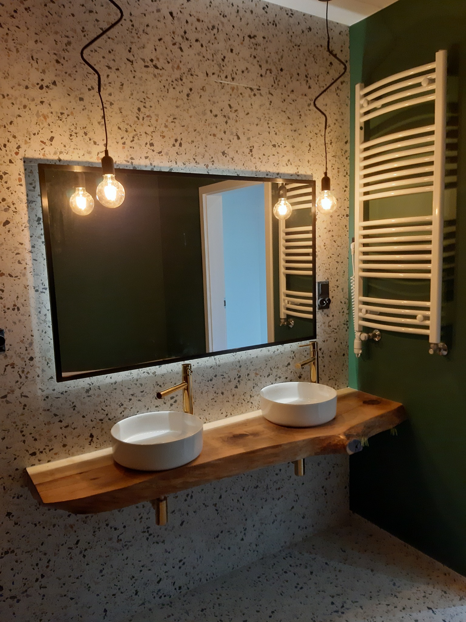 Nowoczesna łazienka z umywalkami nablatowymi na drewnianym blacie, ściany i podłoga w lastryko, minimalistyczne lampy wiszące z żarówkami Edisona, grzejnik drabinkowy na zielonej ścianie.