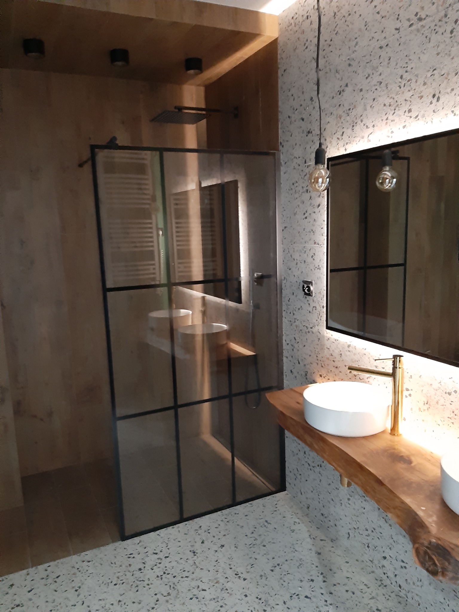 Nowoczesna łazienka z kabiną prysznicową typu walk-in z czarnymi ramami, umywalkami nablatowymi na blacie z surowego drewna, oświetleniem punktowym i lustrem z podświetleniem LED.