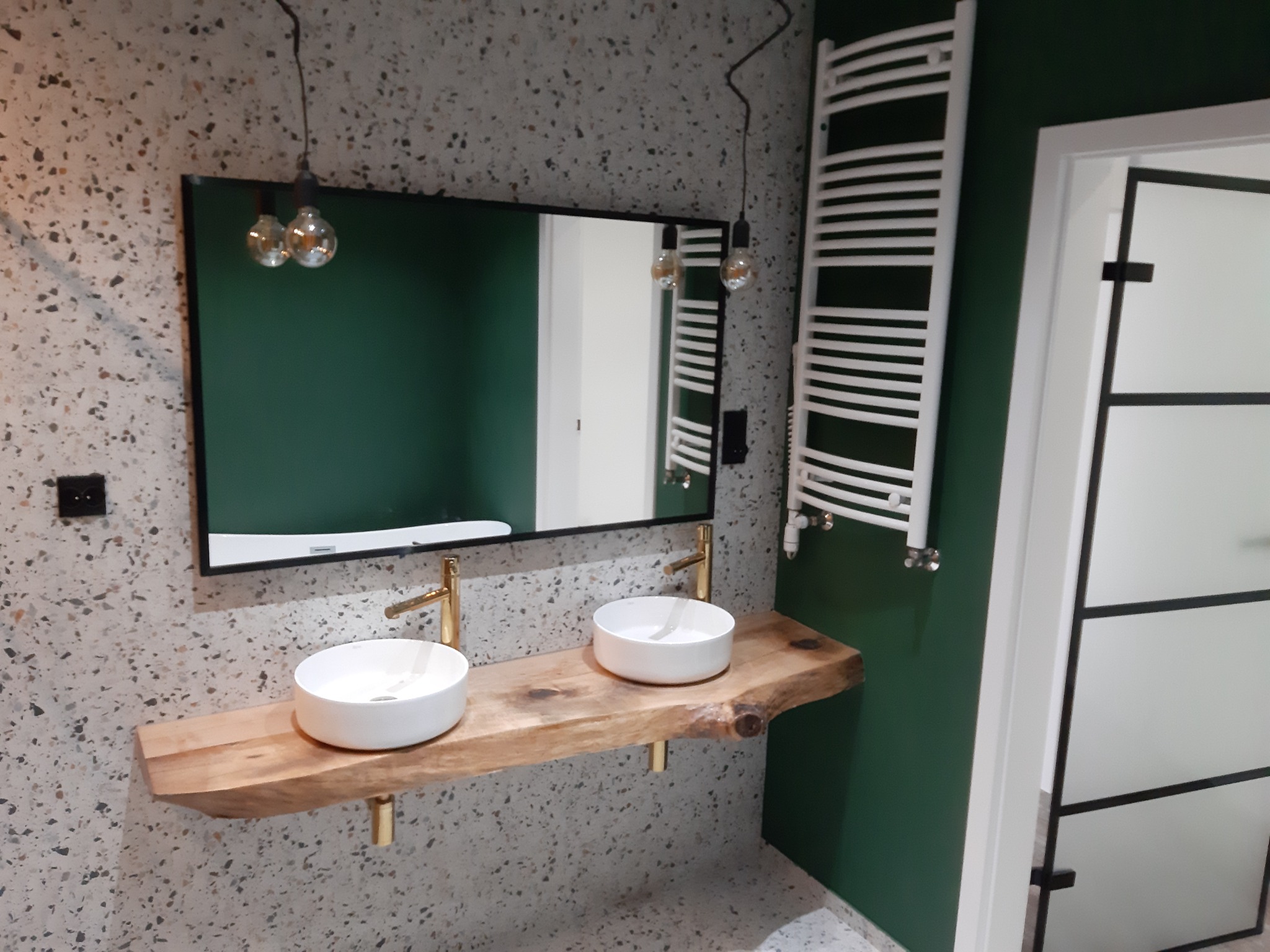 Nowoczesna łazienka z dwoma białymi umywalkami nablatowymi na drewnianym blacie o naturalnych krawędziach, złotymi bateriami, lustrem w czarnej ramie, wiszącym grzejnikiem drabinkowym i ścianami...