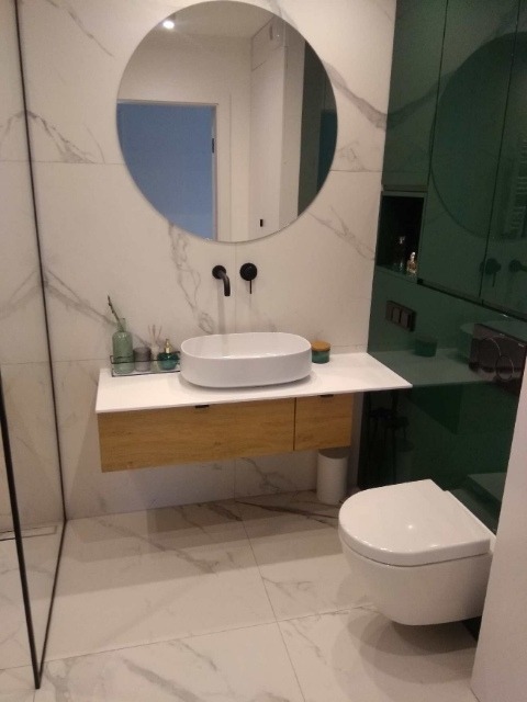 Nowoczesna łazienka z okrągłym lustrem, białą umywalką nablatową na drewnianej szafce, białą toaletą wiszącą i zieloną zabudową meblową, wykończona płytkami imitującymi marmur.