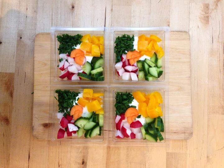 Cztery przezroczyste pudełka z pokrojonymi warzywami (rzodkiewki, ogórki, marchew, papryka, szczypiorek) i sosem na drewnianej desce.