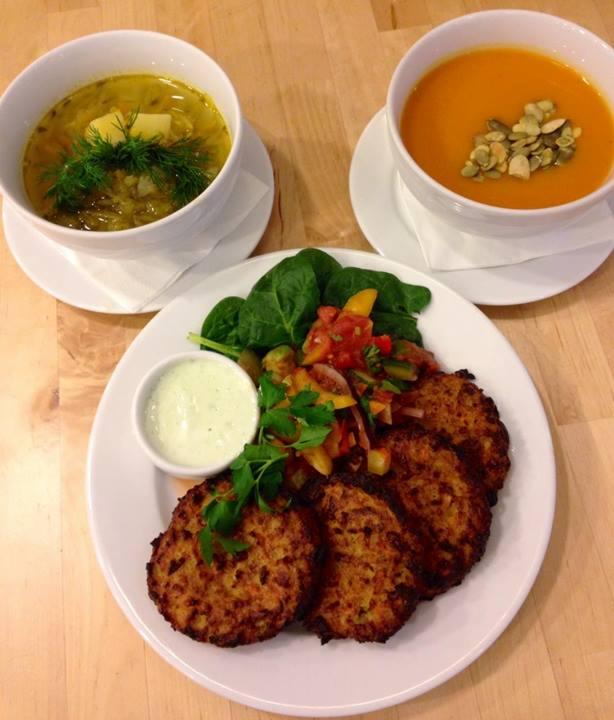 Dwa rodzaje zup w białych miseczkach, jedna z koperkiem i ziemniakami, druga dyniowa z pestkami, oraz talerz z plackami, sałatką ze szpinakiem i salsą warzywną, plus sos ziołowy, na drewnianym stole.