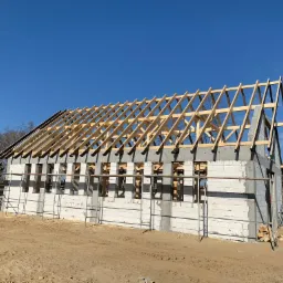 Budowa domu jednorodzinnego w trakcie wznoszenia więźby dachowej; widoczne bloczki betonowe, drewniane elementy konstrukcji i pracownik w niebieskim ubraniu na zewnątrz budynku.