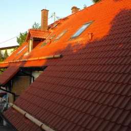 Czerwona dachówka ceramiczna na skośnym dachu z oknami połaciowymi, oświetlona popołudniowym słońcem, widoczny komin i rynny, z boku kontener na gruz i narzędzia.