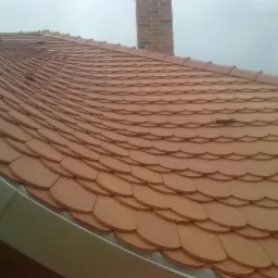 Dach pokryty łuską ceramiczną w odcieniach brązu, widoczny komin z cegły i metalowa obróbka blacharska przy krawędzi dachu.