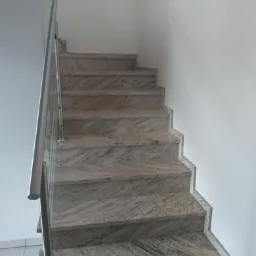 Bieg schodów wewnętrznych wykończonych płytami z jasnego kamienia z wyraźnym wzorem żyłkowania, z metalową balustradą ze szklanym wypełnieniem, widok od dołu.