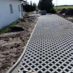 Szara nawierzchnia z ażurowych betonowych płyt ułożona wzdłuż białego budynku, widoczne przygotowane podłoże i paleta płyt w tle.