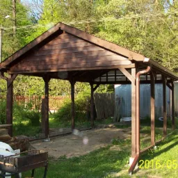 Brązowa, drewniana konstrukcja z rynnami, prawdopodobnie wiata lub zadaszenie, w trakcie budowy, z widocznymi elementami przygotowanymi do montażu, na tle zieleni i zaparkowanego samochodu...