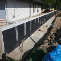 Wzmocniona ściana oporowa z izolacją bitumiczną, widoczna z perspektywy gruntu, z budynkiem w tle i sprzętem budowlanym w oddali.