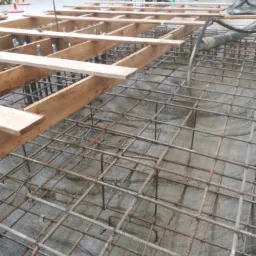 Zalewanie betonem zbrojonej konstrukcji szkieletowej na budowie, widoczne drewniane podpory i wąż do betonu.