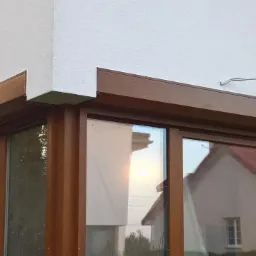 Zamknięta brązowa osłona okienna na elewacji domu z widocznym odbiciem sąsiedniego budynku w oknie.