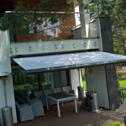 Rozwinięta markiza tarasowa w kolorze szarym, zamontowana na nowoczesnym domu z drewnianym tarasem, widoczne grzejniki tarasowe i meble ogrodowe.