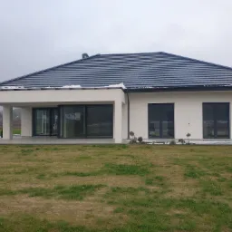 Nowoczesny dom jednorodzinny w Lęborku z antracytową dachówką i dużymi przesuwnymi oknami tarasowymi, widok z zewnątrz na trawnik w pochmurny dzień.