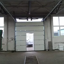 Otwarta segmentowa brama w hali przemysłowej, widoczny fragment samochodu dostawczego w otworze, metalowa konstrukcja sufitu i rampy na posadzce.