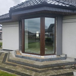 HMB MIKRON - Wystający narożny okap z brązową ramą okienną, widok z zewnątrz na etapie montażu, zabezpieczony folią i uszczelniony pianką montażową, z widocznymi schodami z kostki brukowej prowadzącymi do okna.