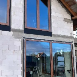 Montaż okien z brązowymi ramami w surowym budynku z białego betonu komórkowego, widoczna drabina i przyssawki do szyb.