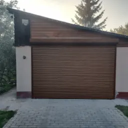 Zewnętrzna brama garażowa rolowana, w brązowym kolorze, zamontowana w budynku z drewnianym szczytem i jasnymi ścianami, widoczne kamery monitoringu.