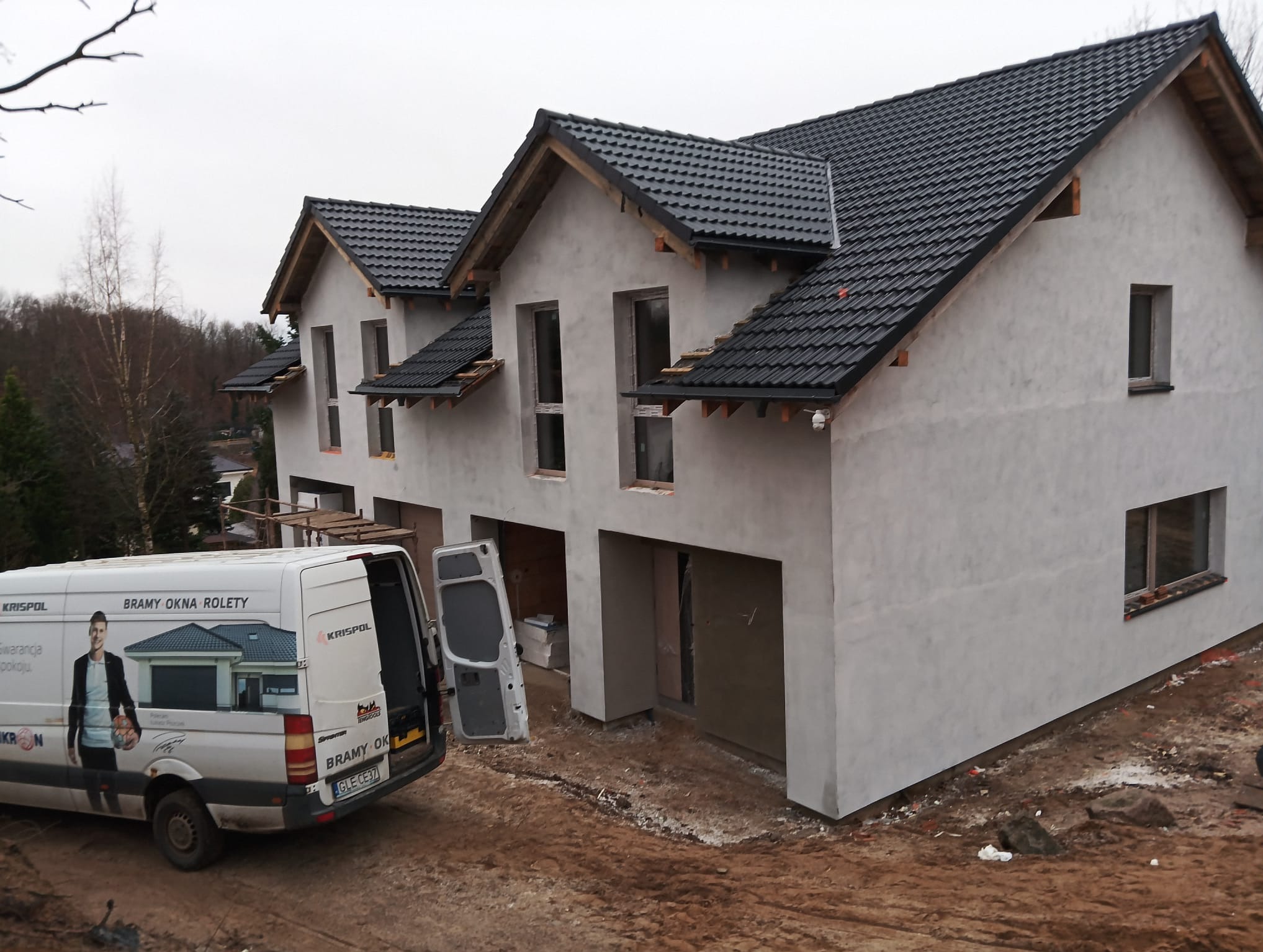 Surowy stan trzypiętrowego domu szeregowego z otworami okiennymi i garażowymi, na pierwszym planie samochód dostawczy z reklamą bram, okien i rolet firmy Krispol.