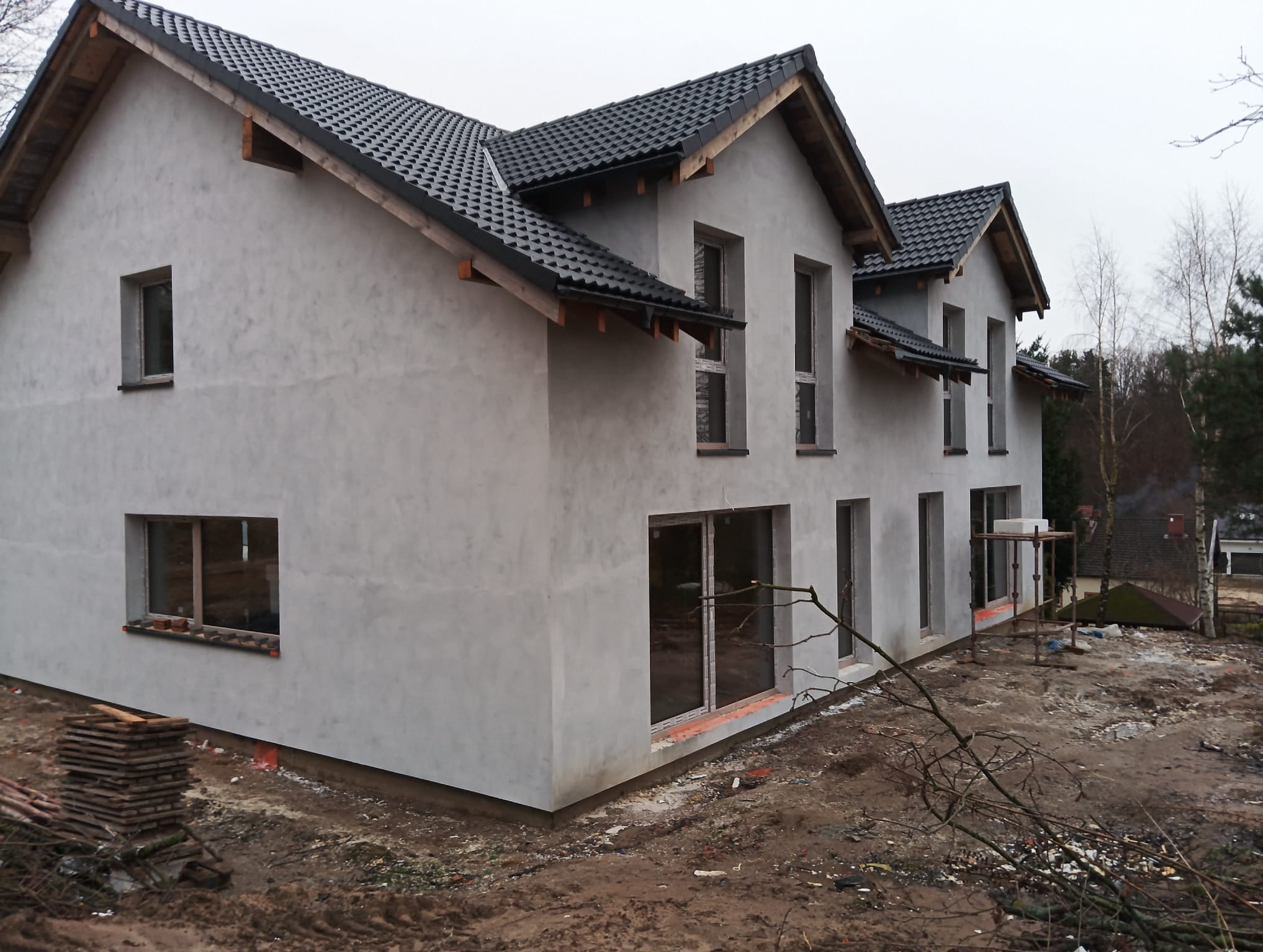 Surowy stan dwukondygnacyjnego domu z szarą elewacją i ciemną dachówką, widoczne otwory okienne w trakcie montażu, teren wokół domu w stanie budowy.