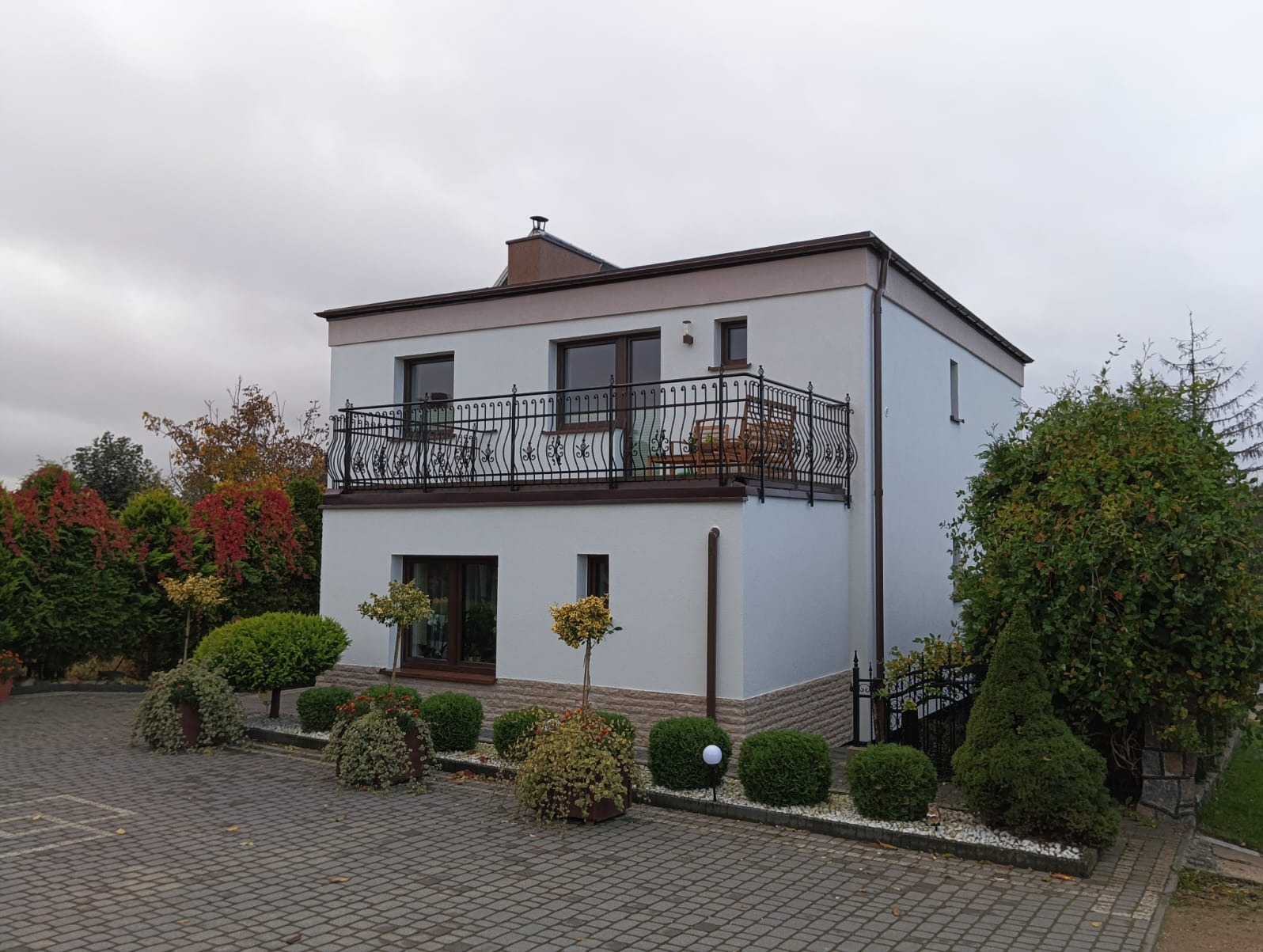 Dwupiętrowy dom z balkonem z kutą, czarną balustradą, brązowe okna i drzwi, kostka brukowa na podjeździe i ozdobne krzewy.
