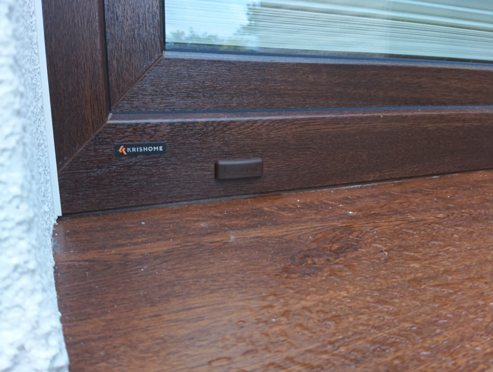 Fragment brązowego okna z imitacją drewna, widoczny zamek i logo producenta KrisHome, przy ścianie o szorstkiej fakturze.