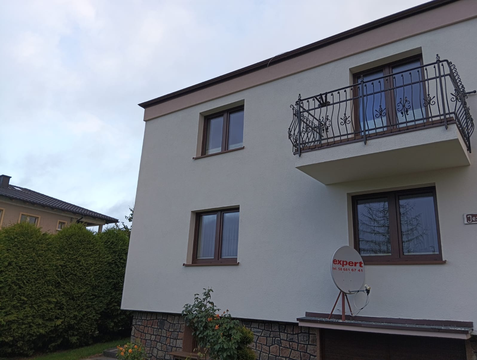 Dwupiętrowy dom z beżową elewacją, brązowymi ramami okiennymi i balkonem z ozdobną, kutą balustradą, widoczny z perspektywy ogrodu z niskim kamiennym murem i roślinnością.