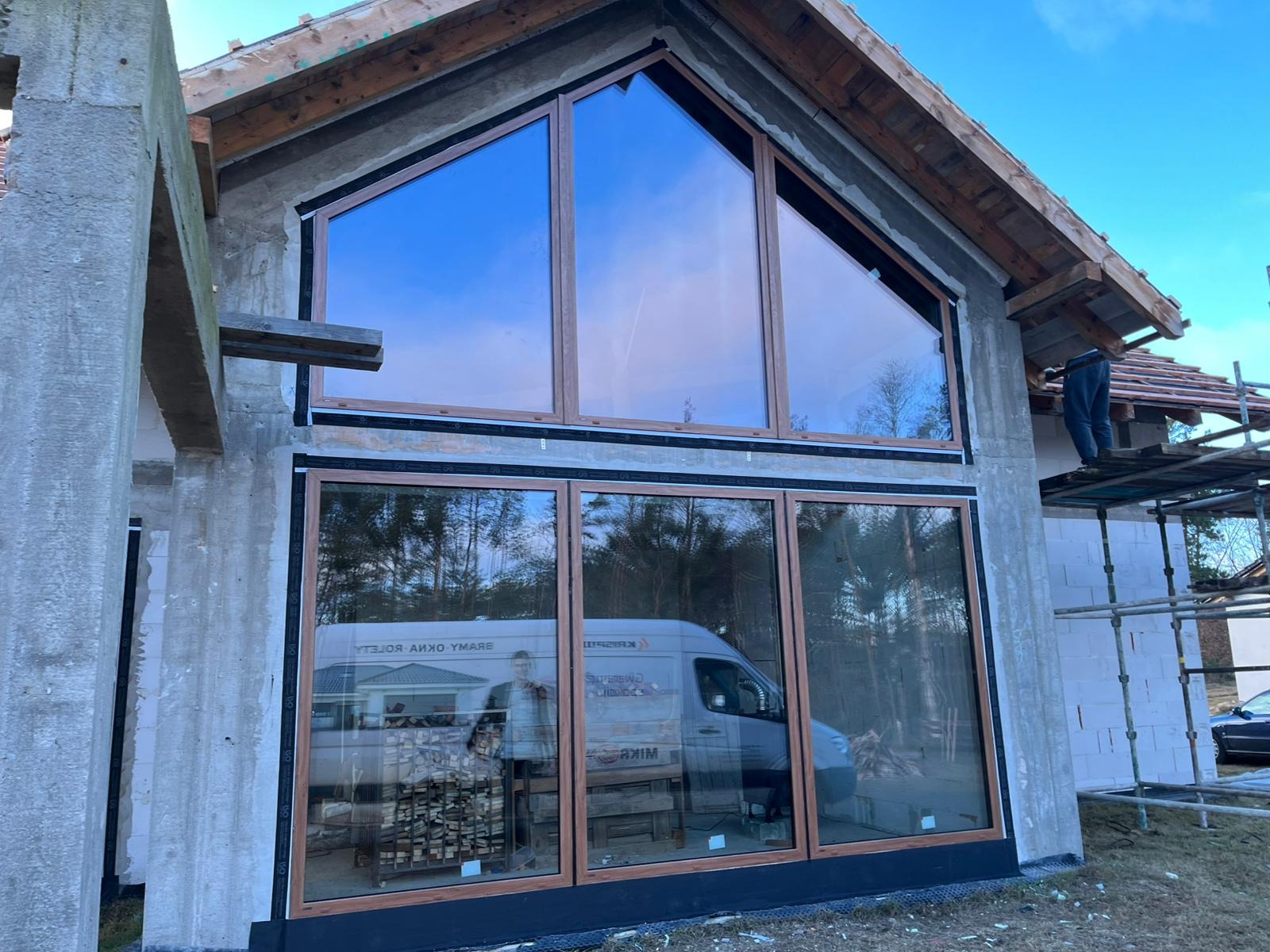 Surowy stan elewacji domu z dużymi oknami z brązowymi ramami, odbicie samochodu dostawczego z pracownikiem w szybach, widoczne rusztowanie i częściowo wykończony dach.